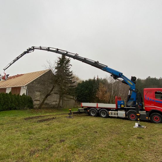 Ein roter Lkw mit einem blauen Kran steht auf einem Rasenfeld und greift nach dem Dach eines Gebäudes.