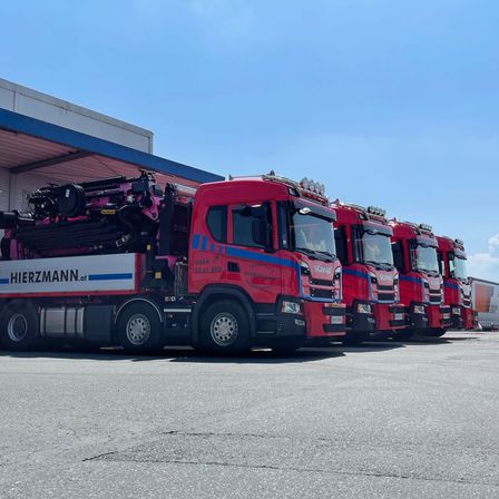 Mehrere rote Scania-Lkw sind in einer Reihe vor einem Gebäude mit blauem Dach geparkt. Die Lkw haben Maschinen daran befestigt.