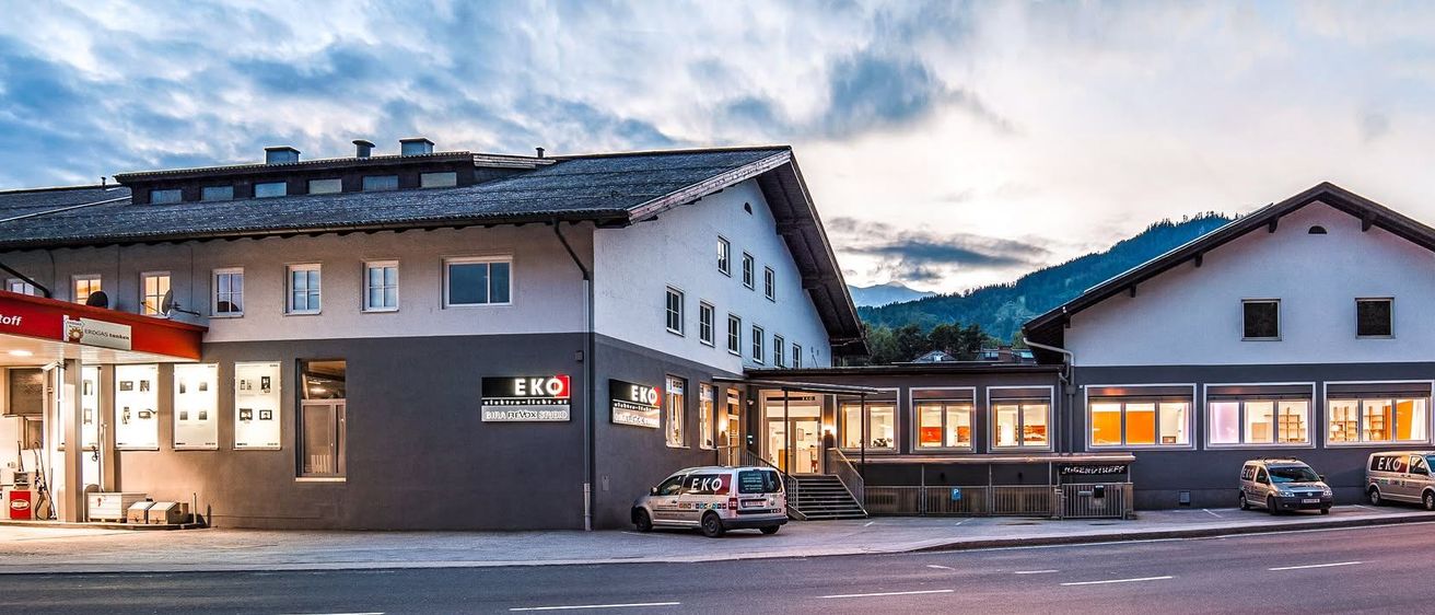 Ein Gebäude mit dem Wort Eko am Schild steht an der Straße. Es hat mehrere Fenster und einen Eingang mit Treppen. Ein Van ist vor dem Gebäude geparkt. Ein Berg ist in der Ferne.