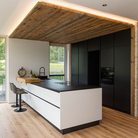 Eine moderne Küche mit Holzdecke, schwarzen Arbeitsplatten und weißen Schränken. Zwei Hocker stehen vor der Arbeitsplatte. Eine Uhr und Pflanzen sind auf der Arbeitsplatte.