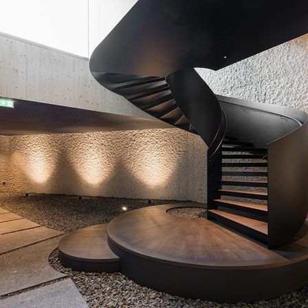 Ein modernes Gebäude verfügt über eine Wendeltreppe mit schwarzen Geländern und Holzstufen, umgeben von einem Kiesbereich und beleuchtet durch Wandleuchten.