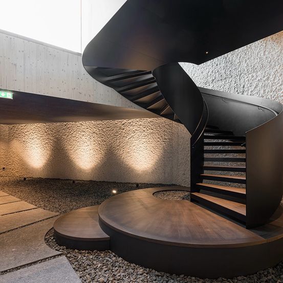 Ein modernes Gebäude verfügt über eine Wendeltreppe mit schwarzen Geländern und Holzstufen, umgeben von einem Kiesbereich und beleuchtet durch Wandleuchten.