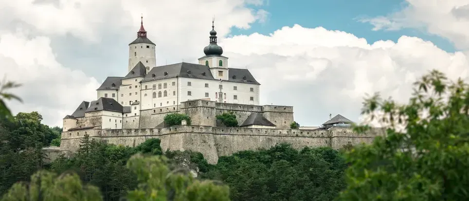 Eine mittelalterliche Burg mit zwei Türmen und einer Kuppel thront auf einem Hügel, umgeben von dichten grünen Bäumen unter einem bewölkten Himmel.