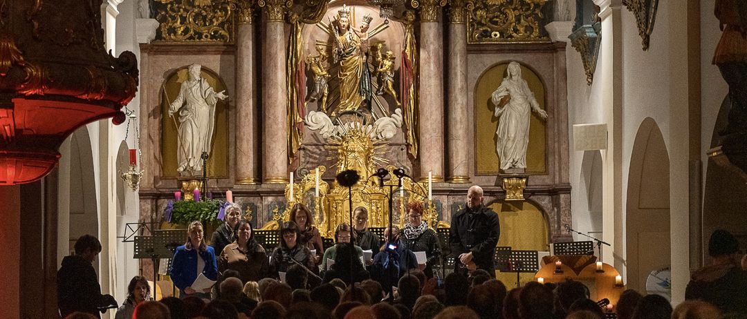 Ein Chor aus Männern und Frauen singt vor einem goldenen Altar in einer Kirche, vor einem großen Publikum. Der Altar ist mit aufwendigen Verzierungen und Statuen von Heiligen geschmückt.