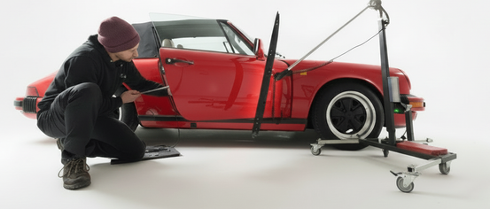 Ein Mann mit Mütze und Turnschuhen kniet neben einem roten Auto, möglicherweise arbeitet er daran. Das Auto steht auf einer weißen Oberfläche, und die Autotür ist geöffnet.
