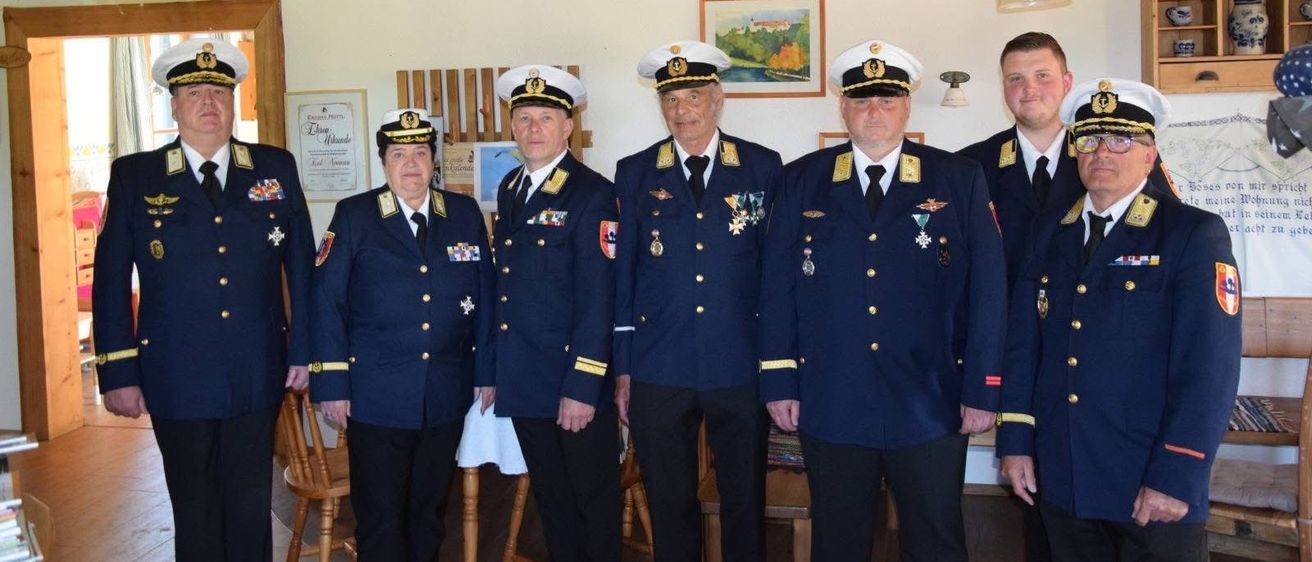Eine Gruppe uniformierter Offiziere steht zusammen, wahrscheinlich in einer militärischen oder polizeilichen Umgebung, in einem Raum mit Holzbalken und einem gerahmten Bild an der Wand.