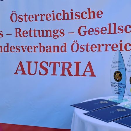 Ein weißer Hintergrund mit rotem Text steht 'Österreichische Rettungs-Geselleschaftsverband Österreich Austria'. Darunter befinden sich drei Auszeichnungen und drei blaue Ordner auf einem Tisch.