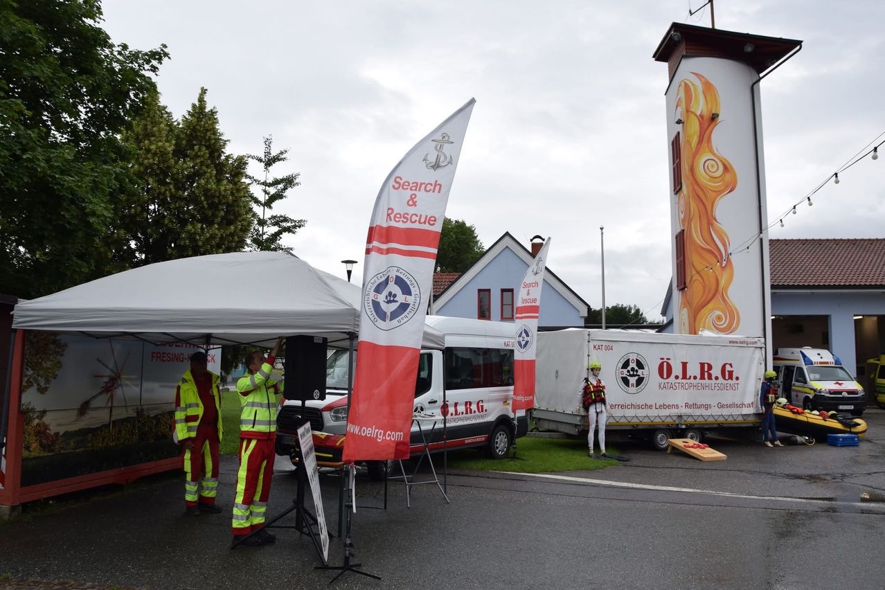 Zwei Personen in Sicherheitsanzügen stehen neben einem Zelt. Ein Banner mit 'Suche & Rettung' ist sichtbar. Dahinter parkt ein weißer Van. Rechts ist ein Anhänger geparkt, gekennzeichnet als 'Katastrophenhilfesdiensten'.