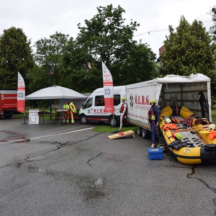 Ein weißer Lieferwagen mit einem roten Banner, das mit 'O.L.R.G' beschriftet ist, ist mit offenem Heck geparkt. Ein gelber Schlauchboot ist im Inneren beladen. Eine Person steht neben dem Boot. Zwei Personen in gelben Westen stehen unter einem Zelt. Bäume und Häuser sind im Hintergrund.