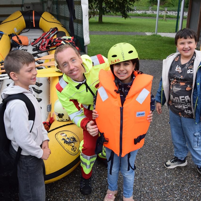 Ein Mann und zwei Kinder stehen neben einem gelben Boot mit Sicherheitsausrüstung. Die Kinder tragen Helme und Rettungswesten.
