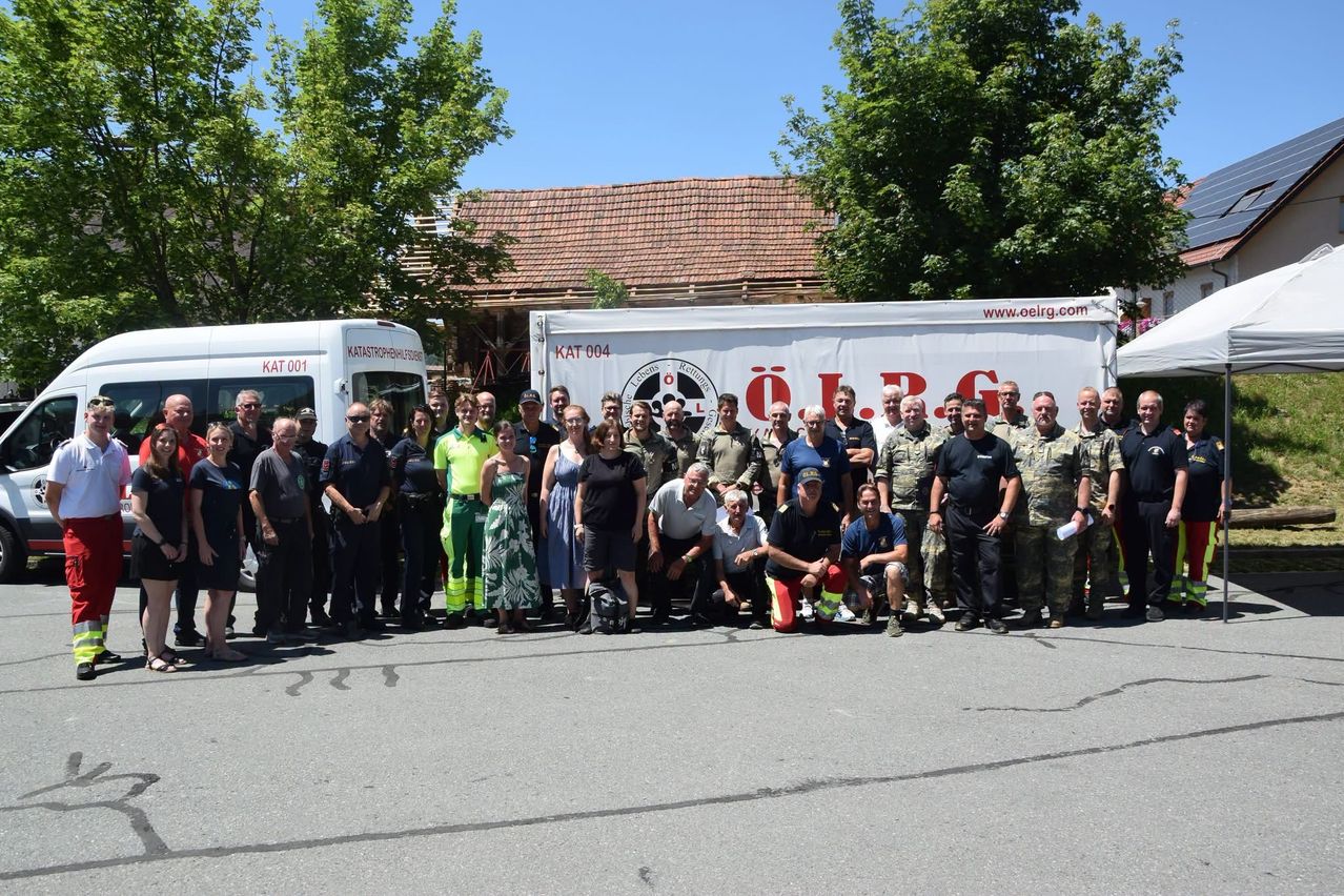 Eine Gruppe von Menschen, einige in Uniformen, posiert für ein Foto vor zwei weißen Fahrzeugen an einem sonnigen Tag. Bäume und ein Haus sind im Hintergrund zu sehen.