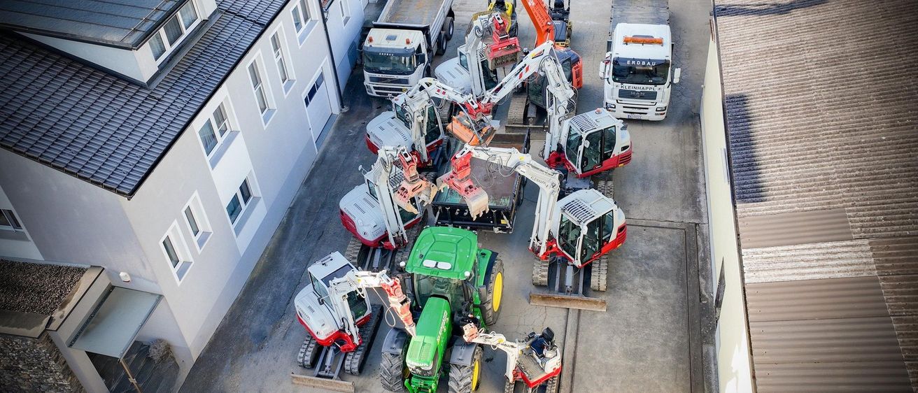 Ein Blick von oben auf eine Baustelle mit mehreren schweren Maschinen geparkt. Darunter sind Kräne, Trucks und Traktoren. Ein weißes Auto ist hinten geparkt.