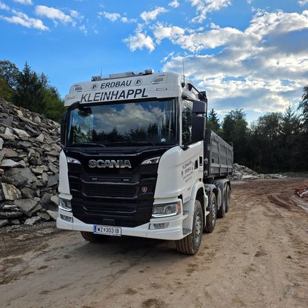 Ein weißer Scania-Lkw mit schwarzem Fahrerhaus fährt auf einer unbefestigten Straße. Der Lkw hat ein Kennzeichen mit der Aufschrift 'WZ 303 IB' und steht neben einem Steinhaufen.