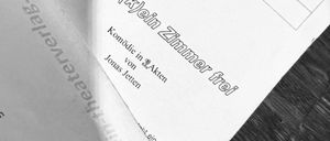 Ein Nahaufnahme eines weißen Papiers mit einem Maskenlogo und schwarzem Text. Das Papier hat einen schwarzen Rand und liegt auf einer Holzoberfläche. Der Text lautet 'In Knast ist (k)jein Zimmer frei.' Das Papier hat auch einen kleinen weißen Maskenaufkleber.