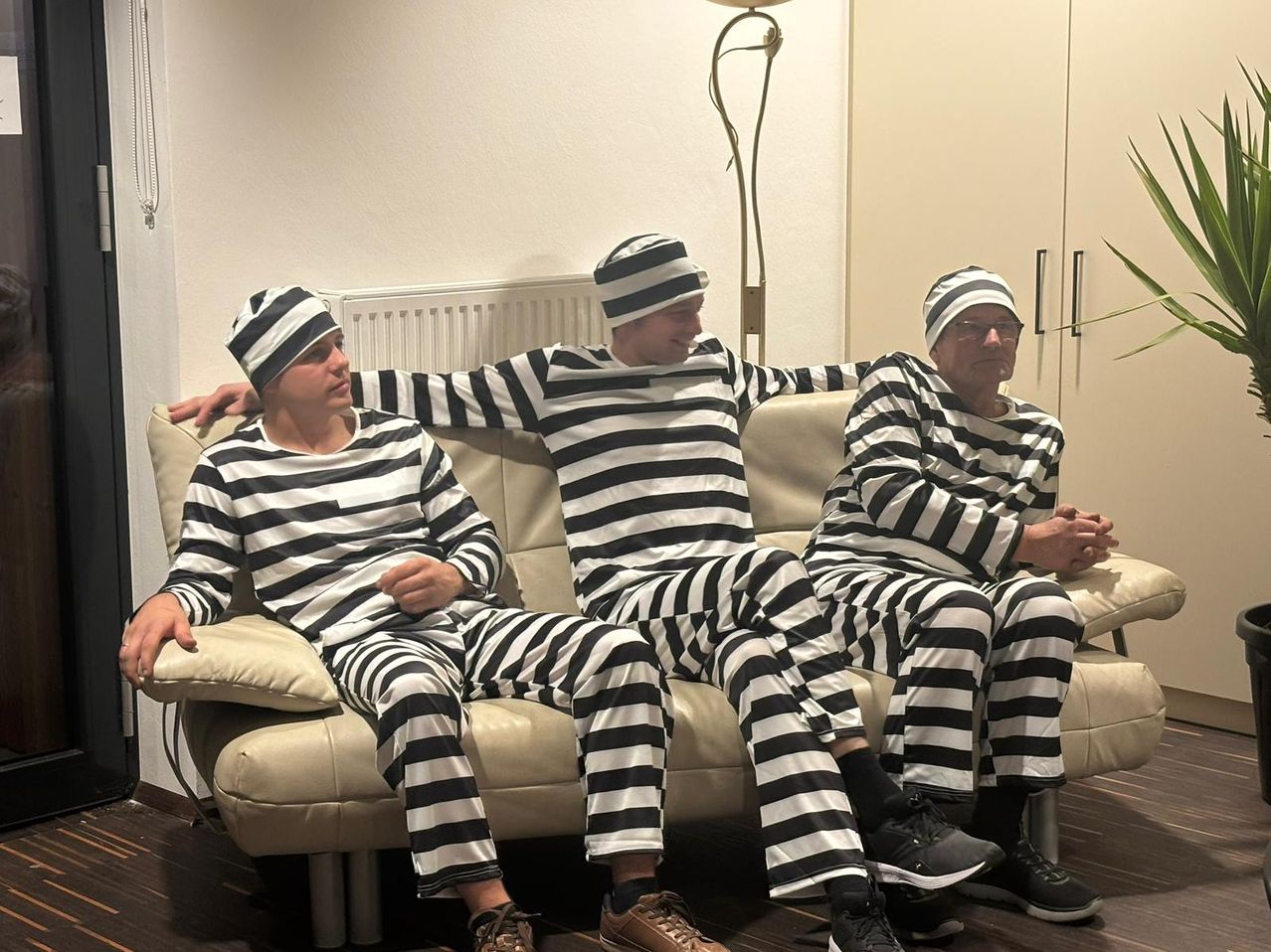 Drei Männer in schwarz-weiß gestreiften Gefängnisuniformen sitzen auf einer Couch. Der Mann auf der linken Seite hat seine Arme um die anderen beiden gelegt.
