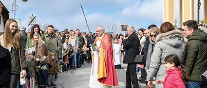 Ein Priester in einer roten und gelben Robe steht vor einer Menschenmenge und hält ein Buch. Die Menge steht am Straßenrand, und einige von ihnen halten Blumen. Es gibt eine Straßenlaterne und ein Gebäude mit einem Kreuz auf der Spitze.