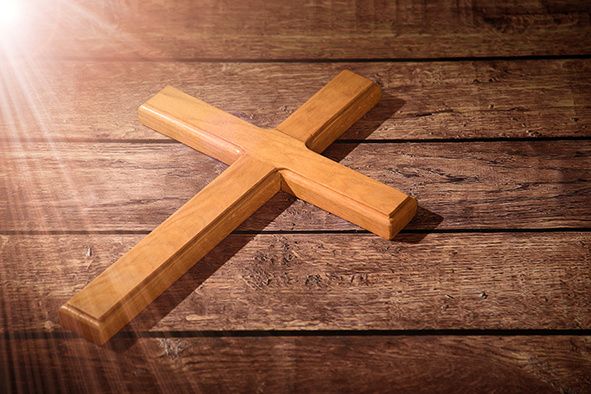 Ein hölzernes Kreuz liegt auf einer Holzoberfläche, wobei das Sonnenlicht von links auf es scheint und einen Schatten auf der rechten Seite wirft.