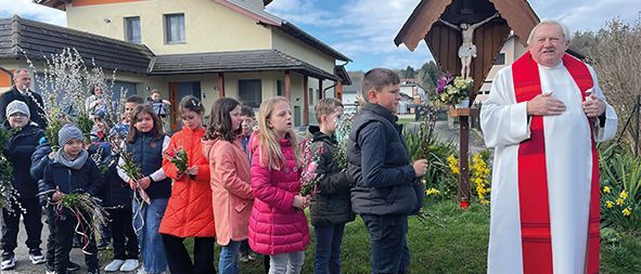 Ein Priester in einer weißen und roten Robe steht vor einem Kruzifix, wobei Kinder Blumen in den Händen halten.