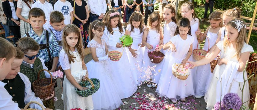 Several children in white dresses are standing in a circle and throwing flower petals. Some are holding baskets with flowers. A crowd of people is standing behind them, watching.