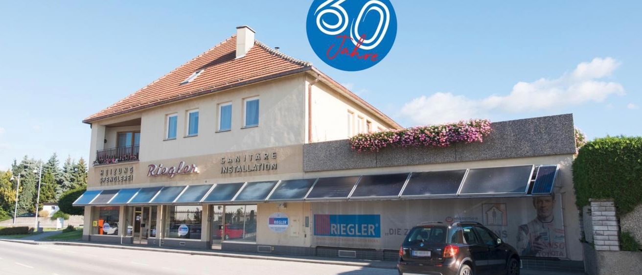 Ein Gebäude mit dem Schild 'Riegler Sanitäre Installation' hat ein 50-Jahre-Logo darüber. Unten am Schild ist ein schwarzes Auto geparkt. Es gibt Blumen an der Wand und Vordächer über den Fenstern.