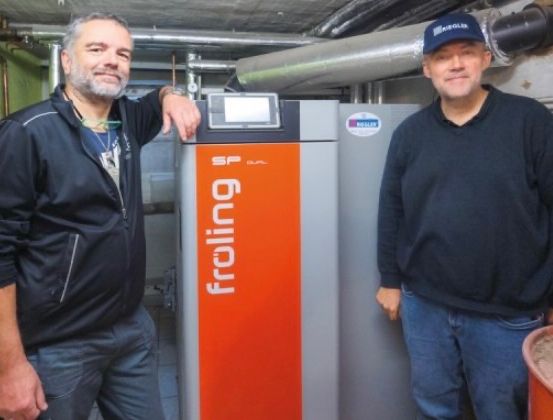 Zwei Männer stehen neben einer großen orange-grauen Maschine mit der Aufschrift 'Froind'. Ein Mann berührt die Maschine, während der andere in die Kamera schaut.
