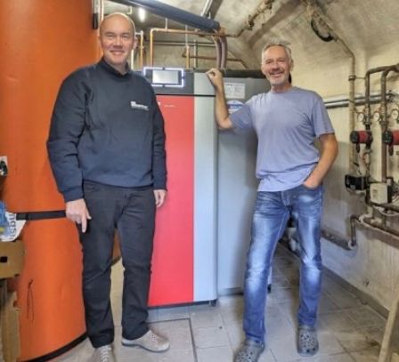 Zwei Männer stehen in einem Raum mit Heizgeräten, lächeln und posieren für ein Foto.