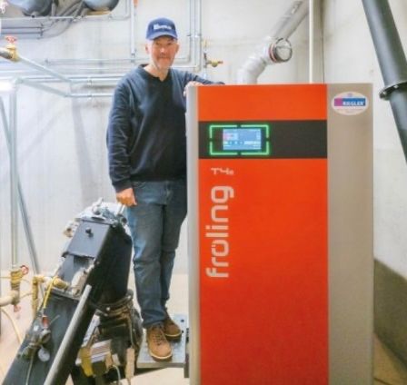 Ein Mann in einem blauen Hut und einem Langarmshirt steht neben einer orangefarbenen und grauen Maschine, möglicherweise einem Heizgerät, mit dem Wort froling darauf.