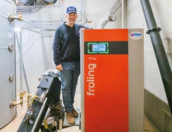 Ein Mann in einem blauen Hut und einem Langarmshirt steht neben einer orangefarbenen und grauen Maschine, möglicherweise einem Heizgerät, mit dem Wort froling darauf.