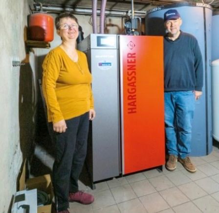 Zwei Personen, ein Mann und eine Frau, stehen in einem Raum mit einem Hargassner-Gerät, blauen und weißen Tanks und Rohren. Sie scheinen für ein Foto zu posieren.