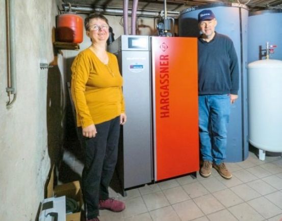 Zwei Personen, ein Mann und eine Frau, stehen in einem Raum mit einem Hargassner-Gerät, blauen und weißen Tanks und Rohren. Sie scheinen für ein Foto zu posieren.