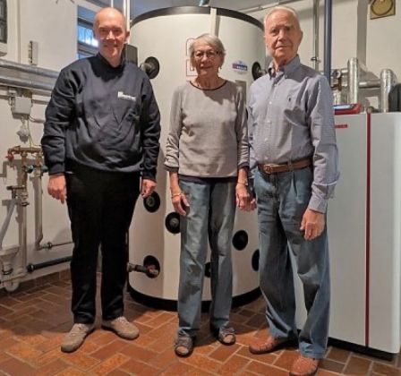 Drei Personen stehen in einem Raum mit Heizgeräten. Der Mann links trägt eine dunkle Jacke, die Frau in der Mitte ein graues Langarmshirt und der Mann rechts ein blaues Hemd.
