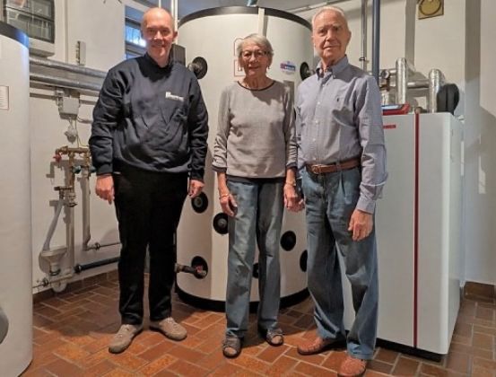 Drei Personen stehen in einem Raum mit Heizgeräten. Der Mann links trägt eine dunkle Jacke, die Frau in der Mitte ein graues Langarmshirt und der Mann rechts ein blaues Hemd.