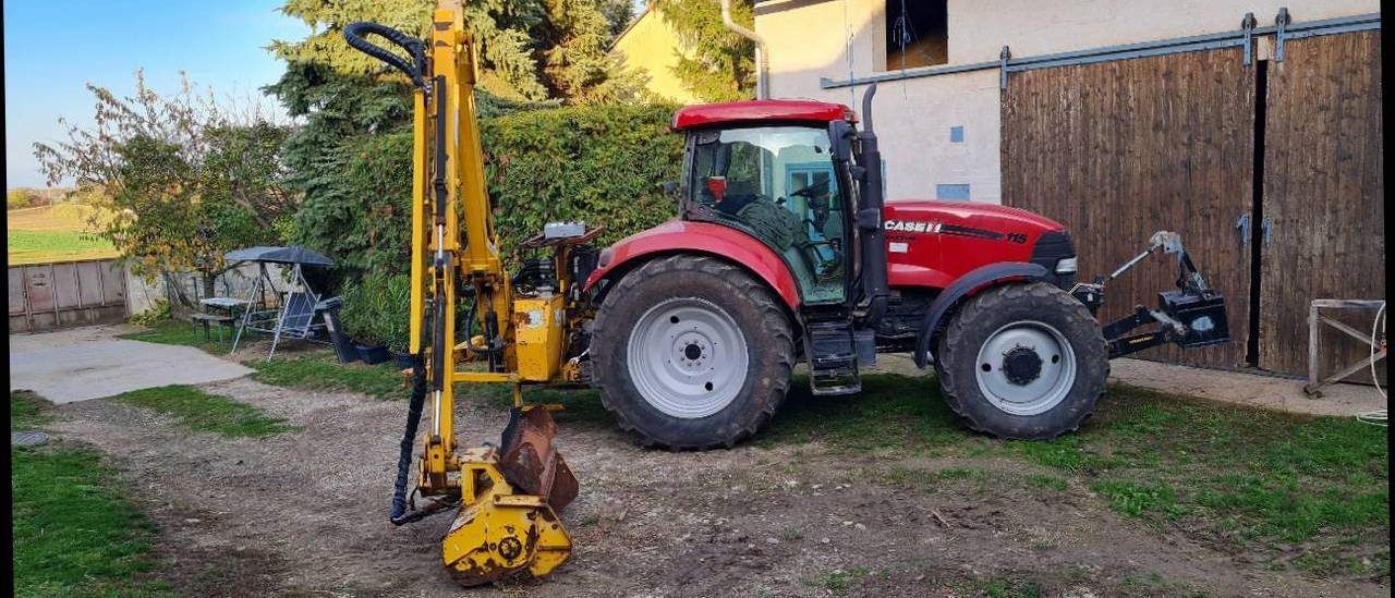 Ein roter Traktor mit einem gelben Anbaugerät steht auf dem Rasen vor einem Haus mit Bäumen und einem Holztor.