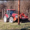 Ein roter Traktor steht auf einem Rasen. Ein Mann sitzt im Inneren und trägt einen Hut. Dahinter steht ein Pfosten und Büsche.