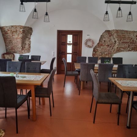 Ein leerer Speisesaal mit Holztischen und Stühlen, Ziegelwänden und Lampen an der Decke. Eine Tür befindet sich in der Mitte, und ein Kranz schmückt die Wand.