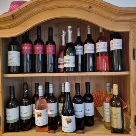 Ein hölzerner Schrank mit zwei Regalen voller Weinflaschen. Das obere Regal hat Miller-Weine und das untere Regal hat Anderl- und Burg-Weine.