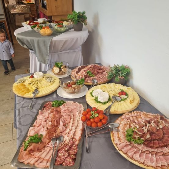 Ein langer Tisch ist mit verschiedenen Fleischsorten, Käse und Gemüse gedeckt. Ein kleines Kind steht in der Nähe. Im Hintergrund befindet sich ein Schrank und eine Wand mit Pflanzen.