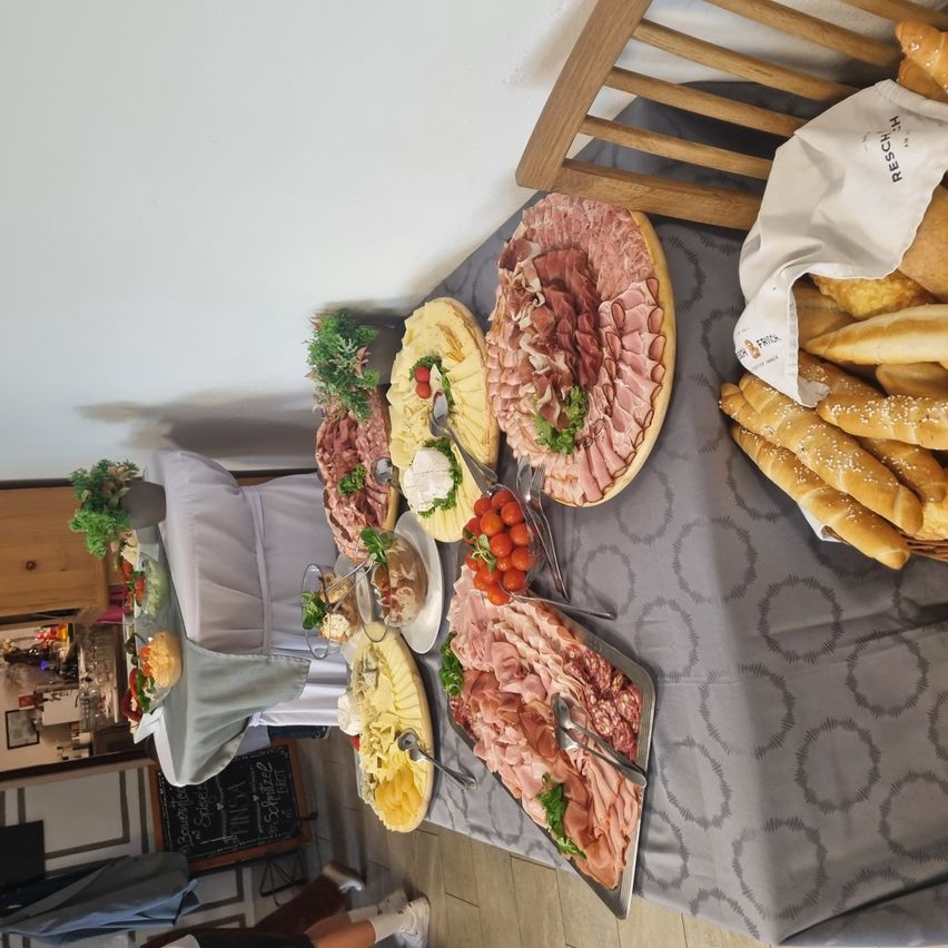 Ein Tisch ist mit einem grauen Tuch bedeckt und hält verschiedene Lebensmittel, darunter Fleisch, Käse und Tomaten. Brot und andere Gegenstände befinden sich an der Seite. Der Hintergrund zeigt eine Holzwand und eine Tafel mit Text.