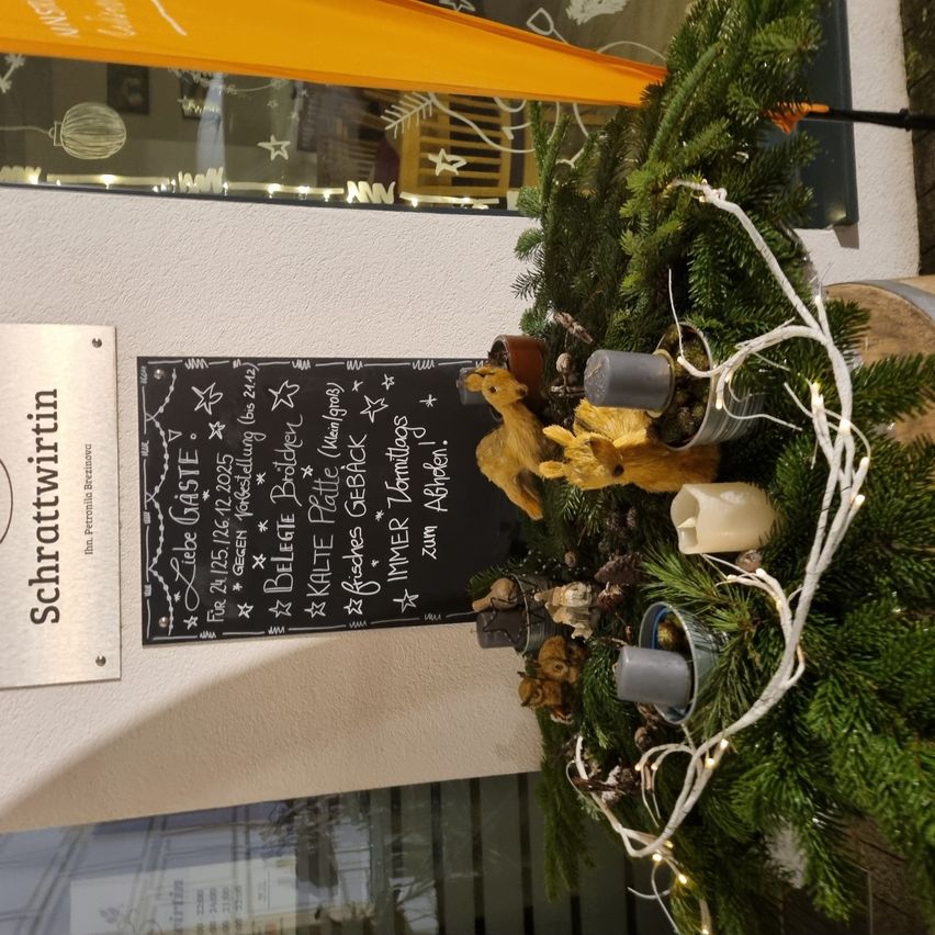Ein dekorativer Kranz mit einer Tafel für das Menü ist an einer Wand befestigt. Der Kranz hat Tannennadeln, weiße Lichter und verschiedene Dekorationen. Die Tafel mit dem Menü listet Speisen und Getränke auf.