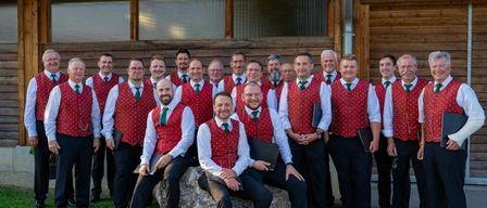 Eine Gruppe von Männern in roten Westen und grünen Krawatten posiert für ein Foto vor einem Holzgebäude.
