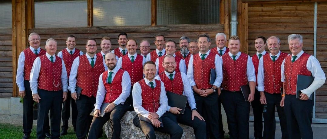 Eine Gruppe von Männern in roten Westen und grünen Krawatten posiert für ein Foto vor einem Holzgebäude.
