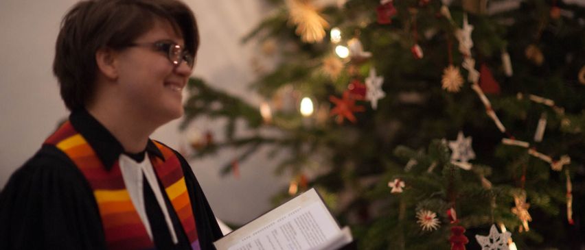 Eine Person in einer Amtstracht und mit Brille hält ein Buch vor einem geschmückten Weihnachtsbaum.