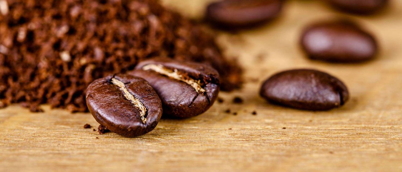 Eine Nahaufnahme von gemahlenen Kaffeebohnen auf einer Holzoberfläche, mit einigen ganzen Bohnen verstreut. Die Bohnen sind dunkelbraun, und einige haben einen sichtbaren Riss.