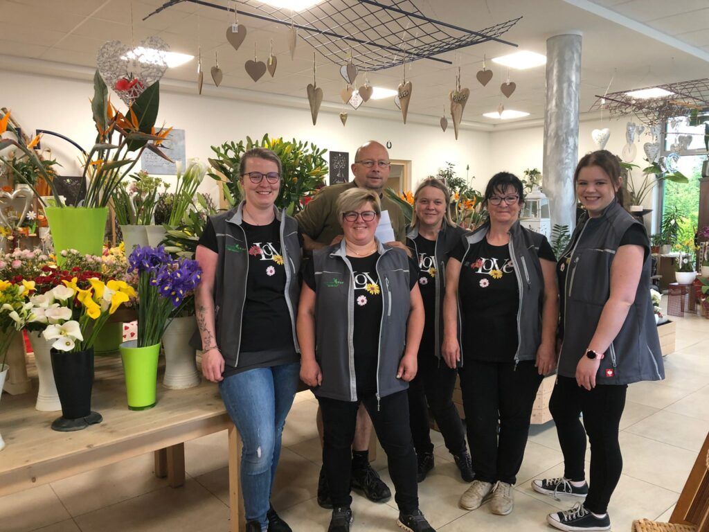 Eine Gruppe von Floristen, darunter Frauen und ein Mann, steht in einem Blumenladen und lächelt für ein Foto, mit verschiedenen Blumen und Herzdekorationen im Hintergrund.