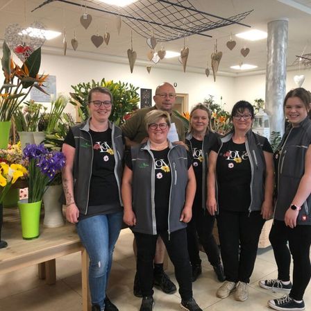 Eine Gruppe von Floristen, darunter Frauen und ein Mann, steht in einem Blumenladen und lächelt für ein Foto, mit verschiedenen Blumen und Herzdekorationen im Hintergrund.