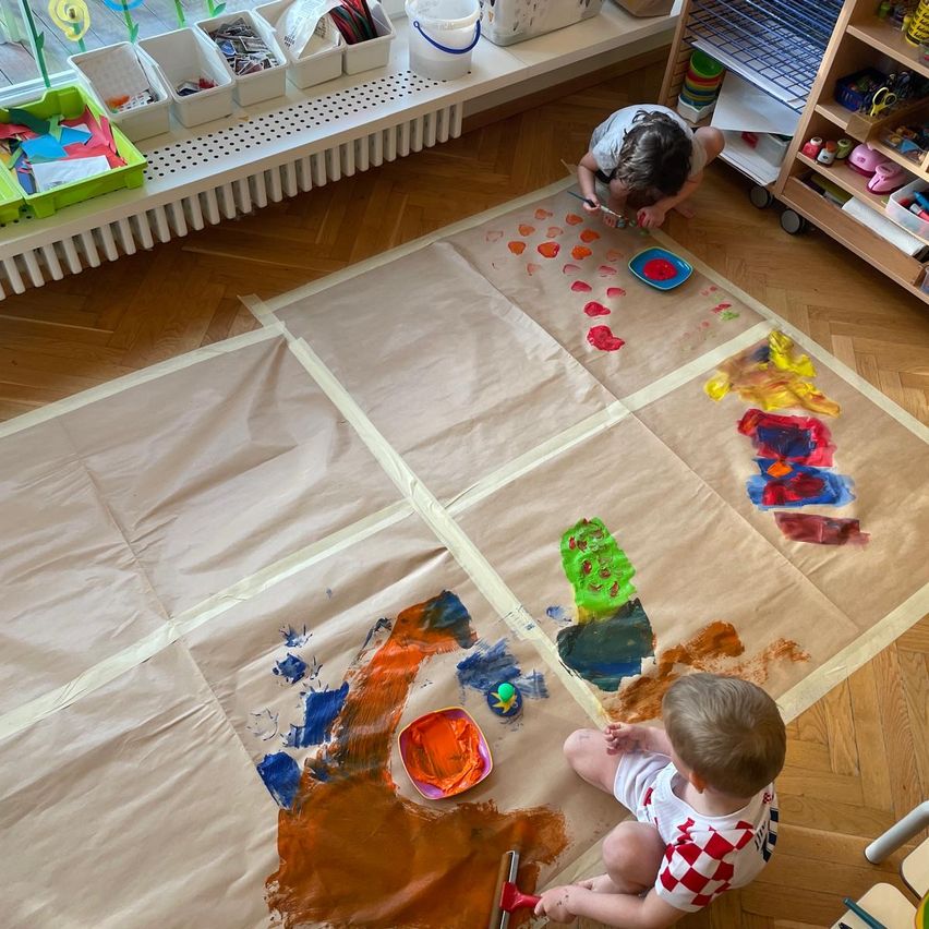 Two young children are painting on a large piece of brown paper laid out on the floor. One child is sitting while the other is kneeling. Various paint colors are visible.