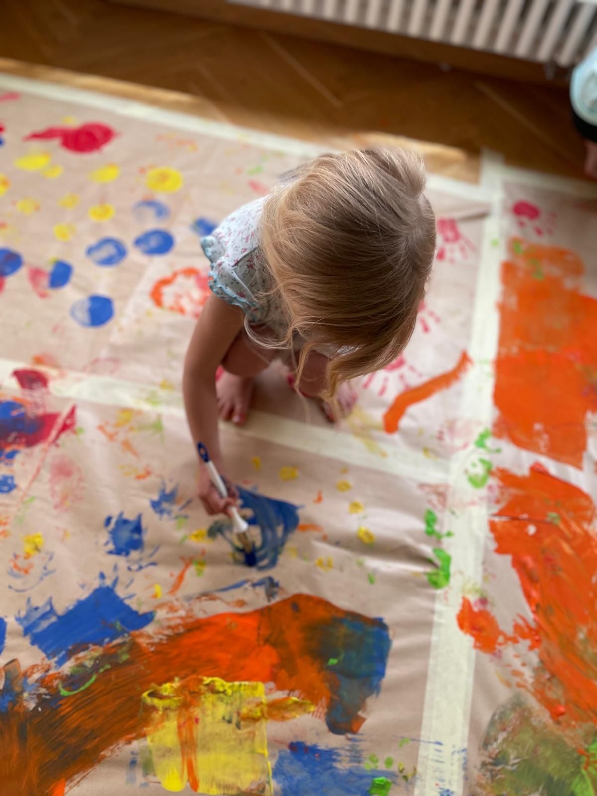 A small child with blonde hair is painting on a large canvas with various colors of paint. The child is holding a paintbrush in her right hand.