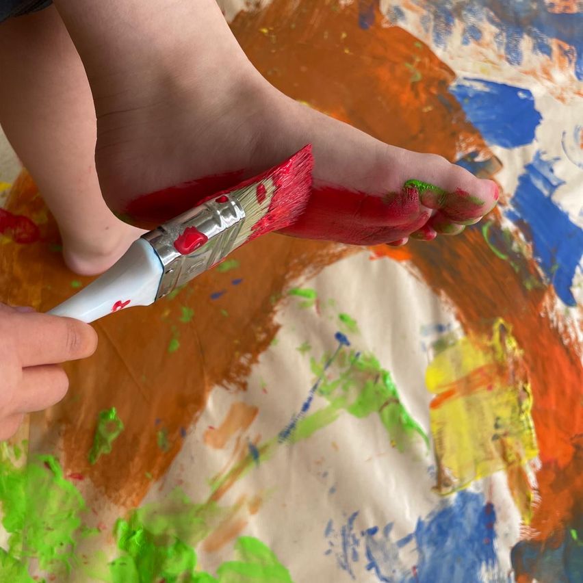 A child is painting with a brush on a canvas covered with various colors of paint. Their feet are on the canvas, with red and green paint on them.