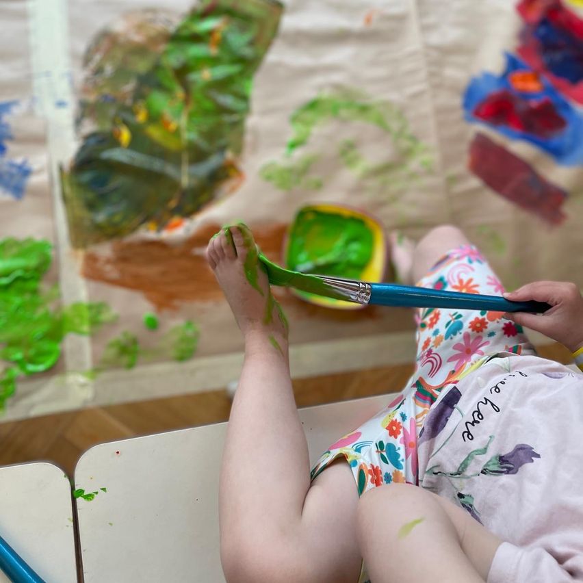 A child is sitting and painting with green paint on their foot on a large canvas. The child has floral pants and a bracelet.