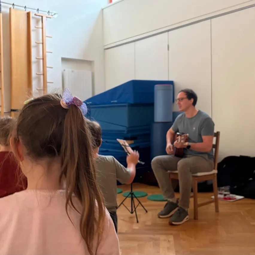 Ein Mann spielt Gitarre, während Kinder zuhören. Er sitzt auf einem Stuhl in einem Raum mit blauen Kissen. Ein Kind hält eine Flöte in der Nähe eines Stativs.